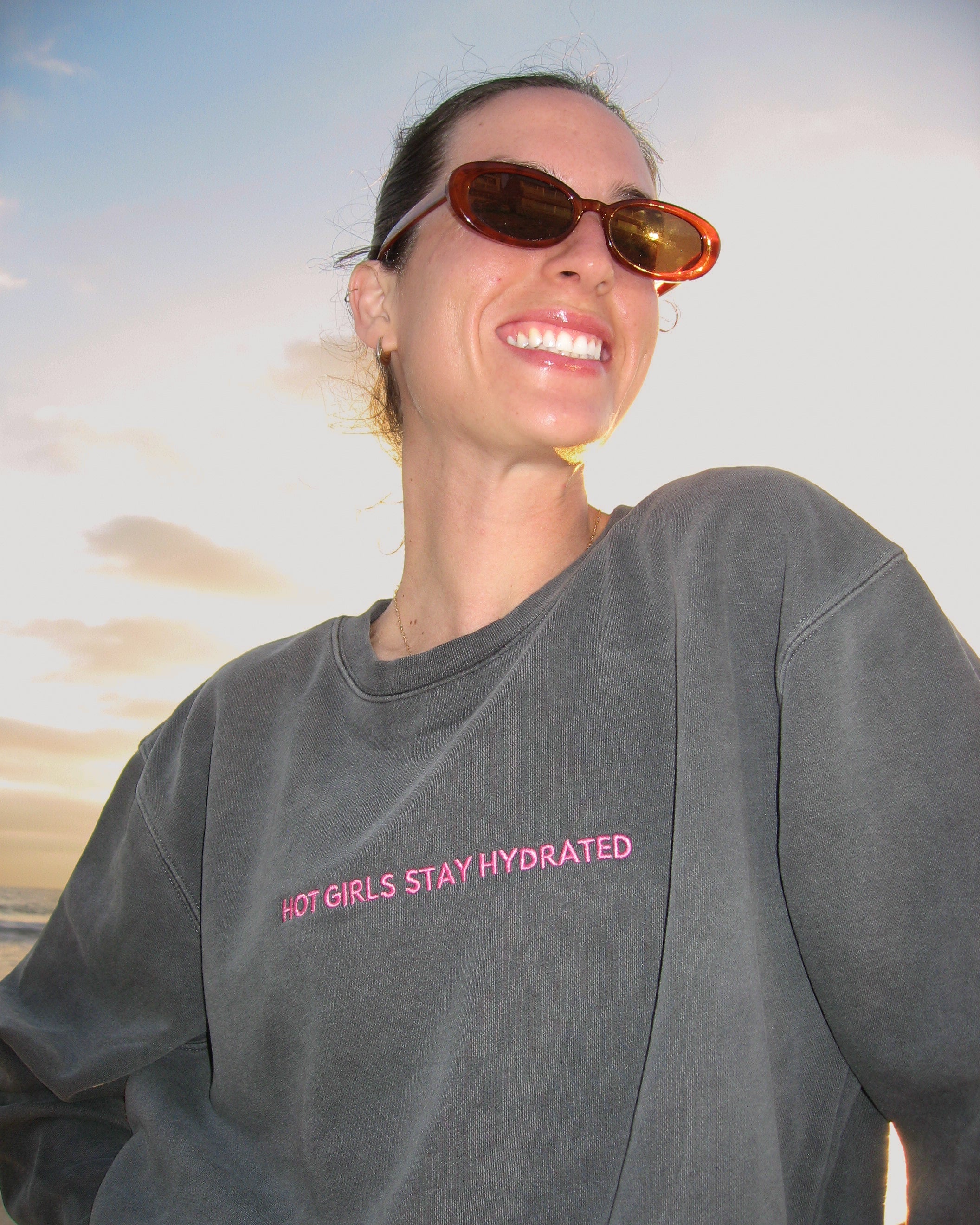 Women wearing sunglasses and a black crewneck with text that says "hot girls stay hydrated" standing against a beach backdrop at sunset.