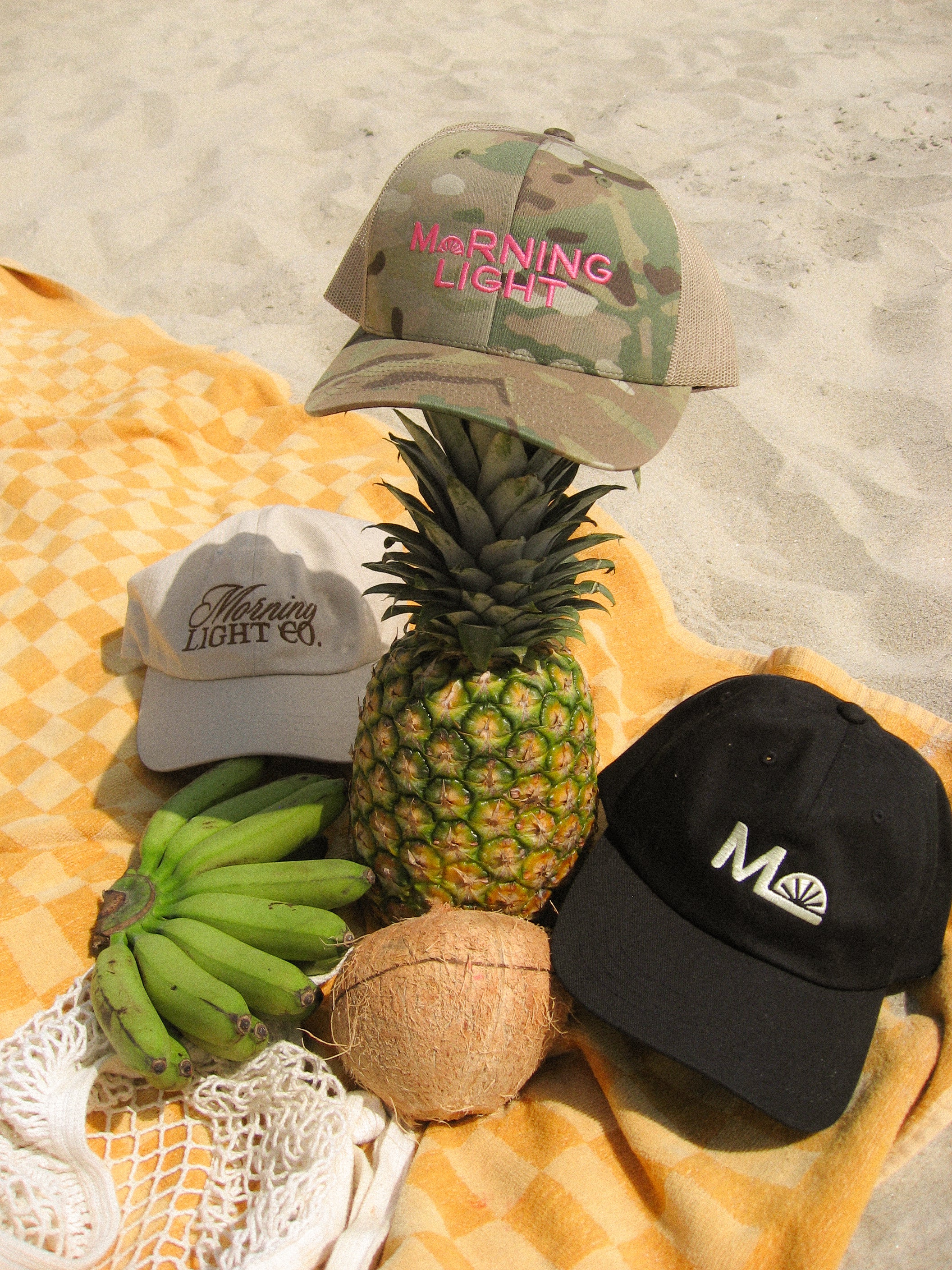 Three baseball caps with 'Morning Light' branding on a beach setting with a pineapple, coconuts, and bananas.