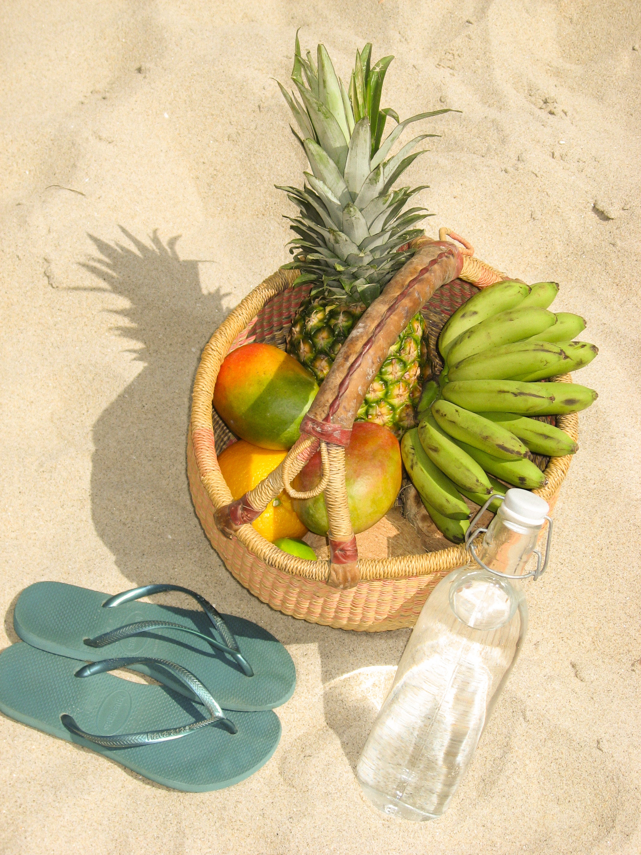 Fruit basket with pineapple, mangoes, and bananas on sandy ground with flip-flops and a bottle.