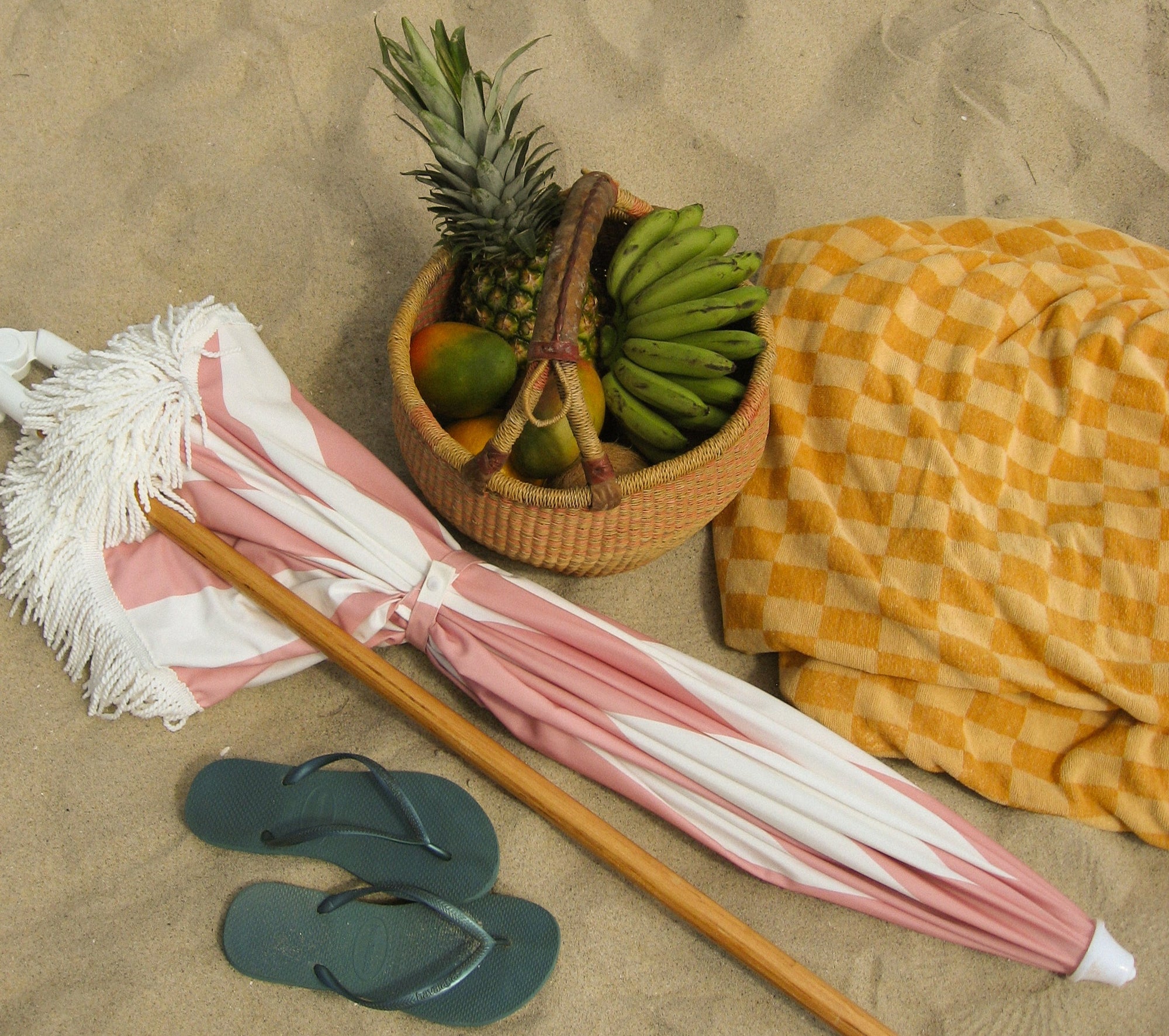 Basket of fruits, beach towel, sandals, and umbrella on sandy beach