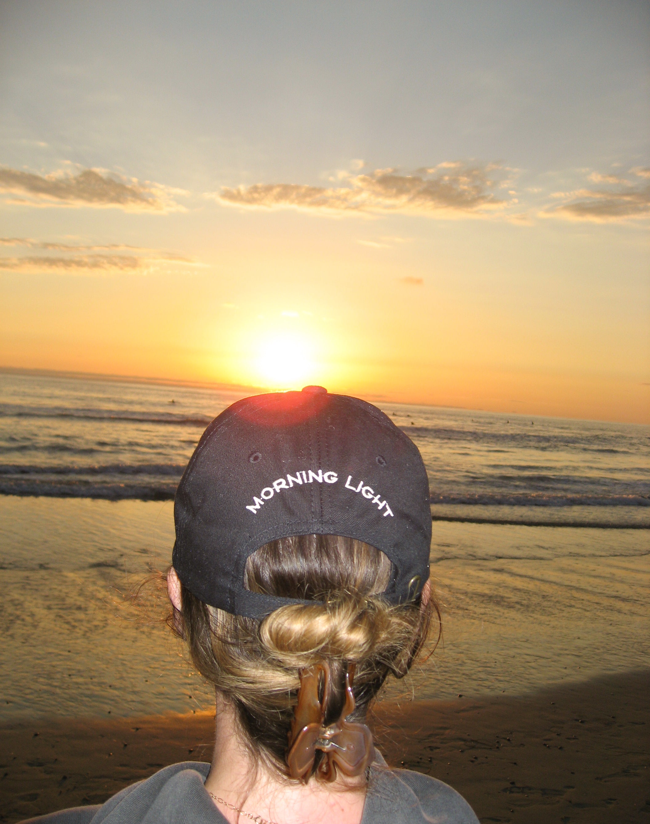 Person wearing a 'Morning Light' cap at sunset on a beach
