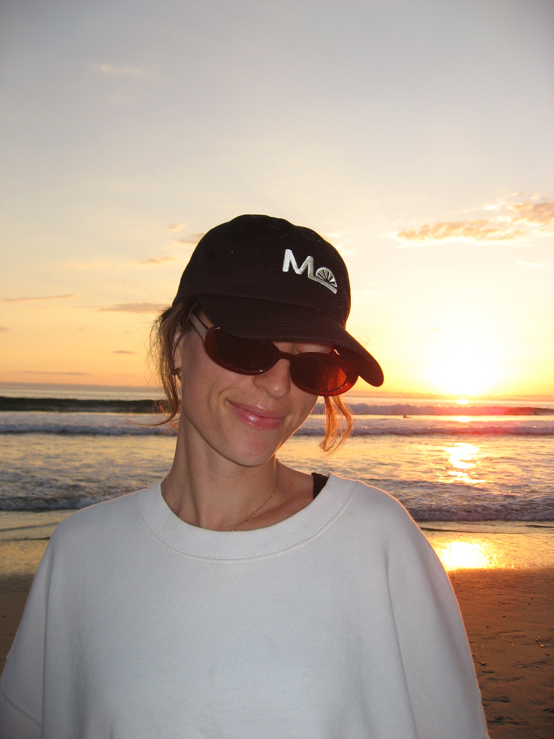 Person wearing a Morning Light cap and sunglasses on a beach at sunset