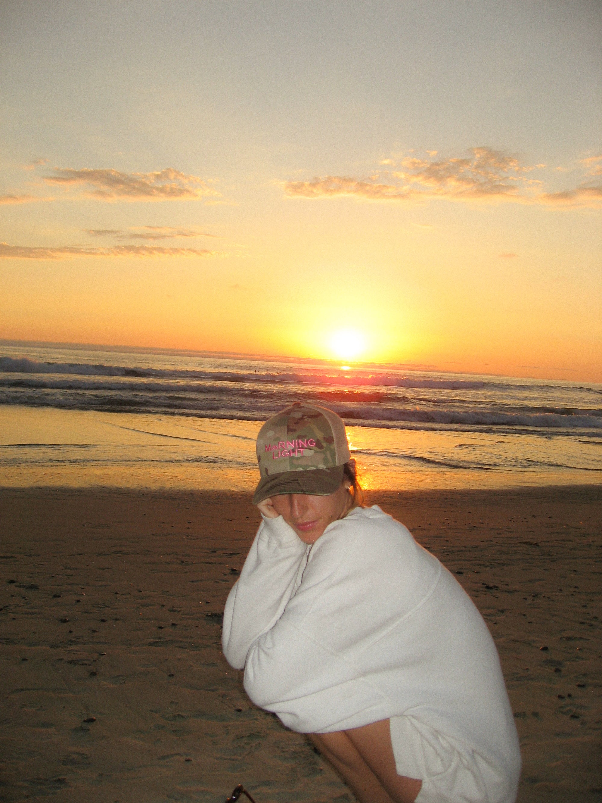 Person sitting down in a white jacket on a beach at sunset wearing a camouflage hat that reads "Morning Light" 