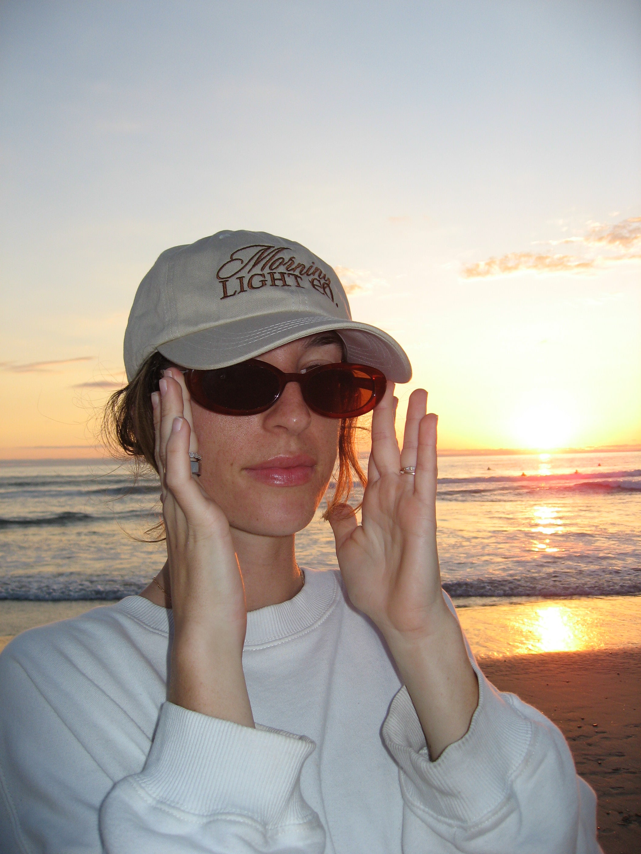 Person wearing a 'Morning Light Co.' cap and sunglasses on a beach at sunset.