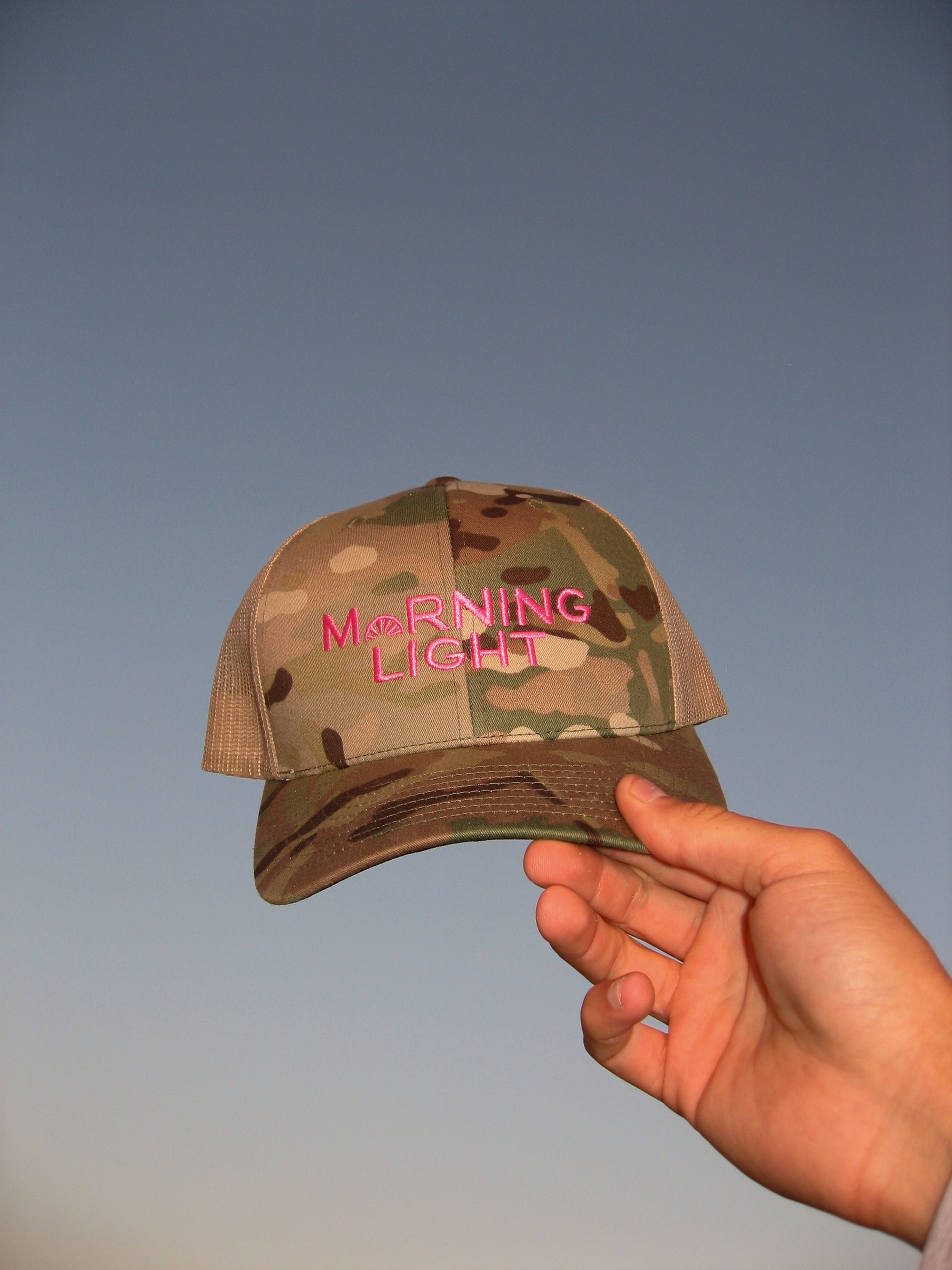 Camouflage cap with 'Morning Light' text held against a gradient blue background