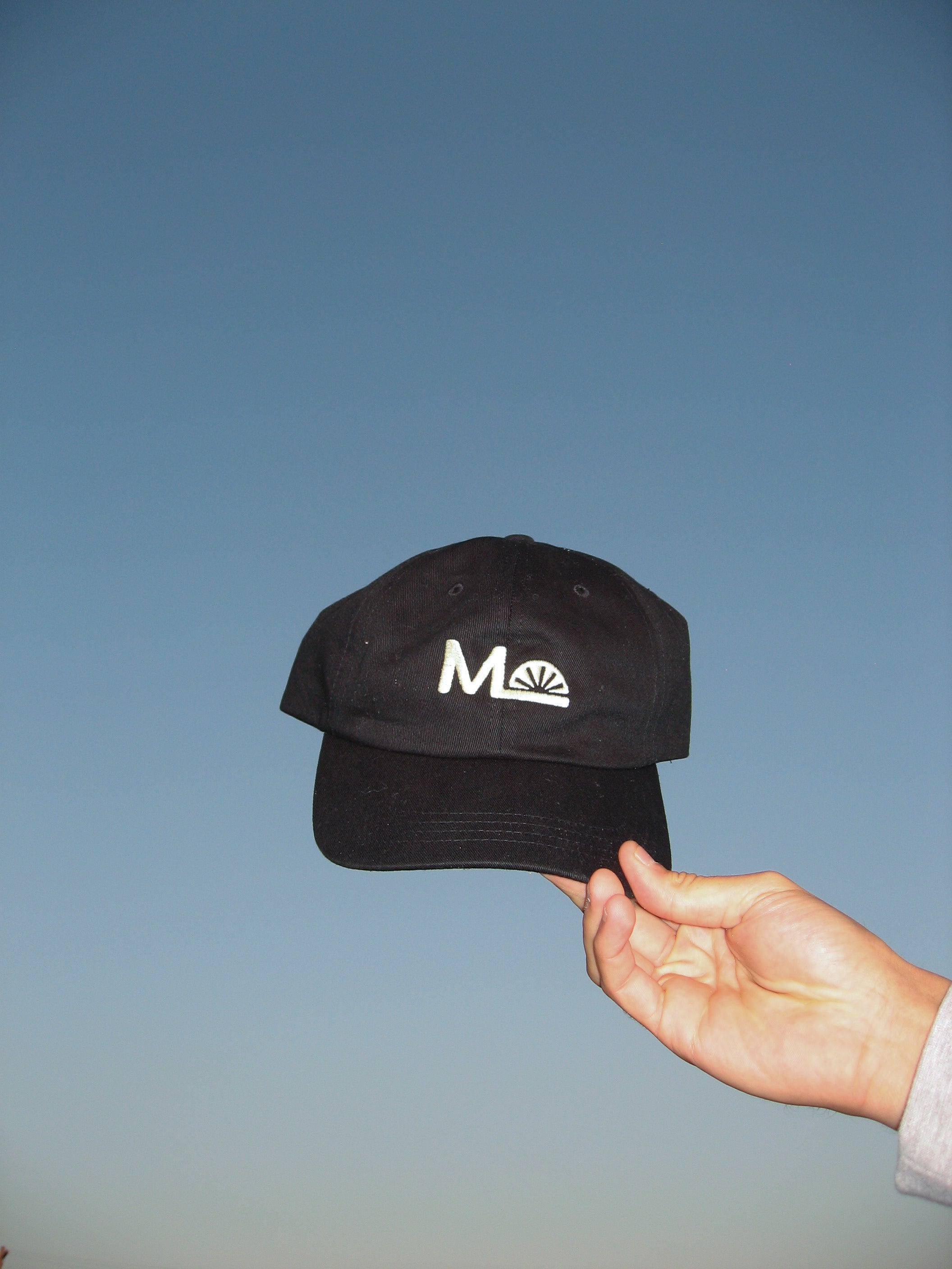 Black cap with a logo held by a hand against a blue sky