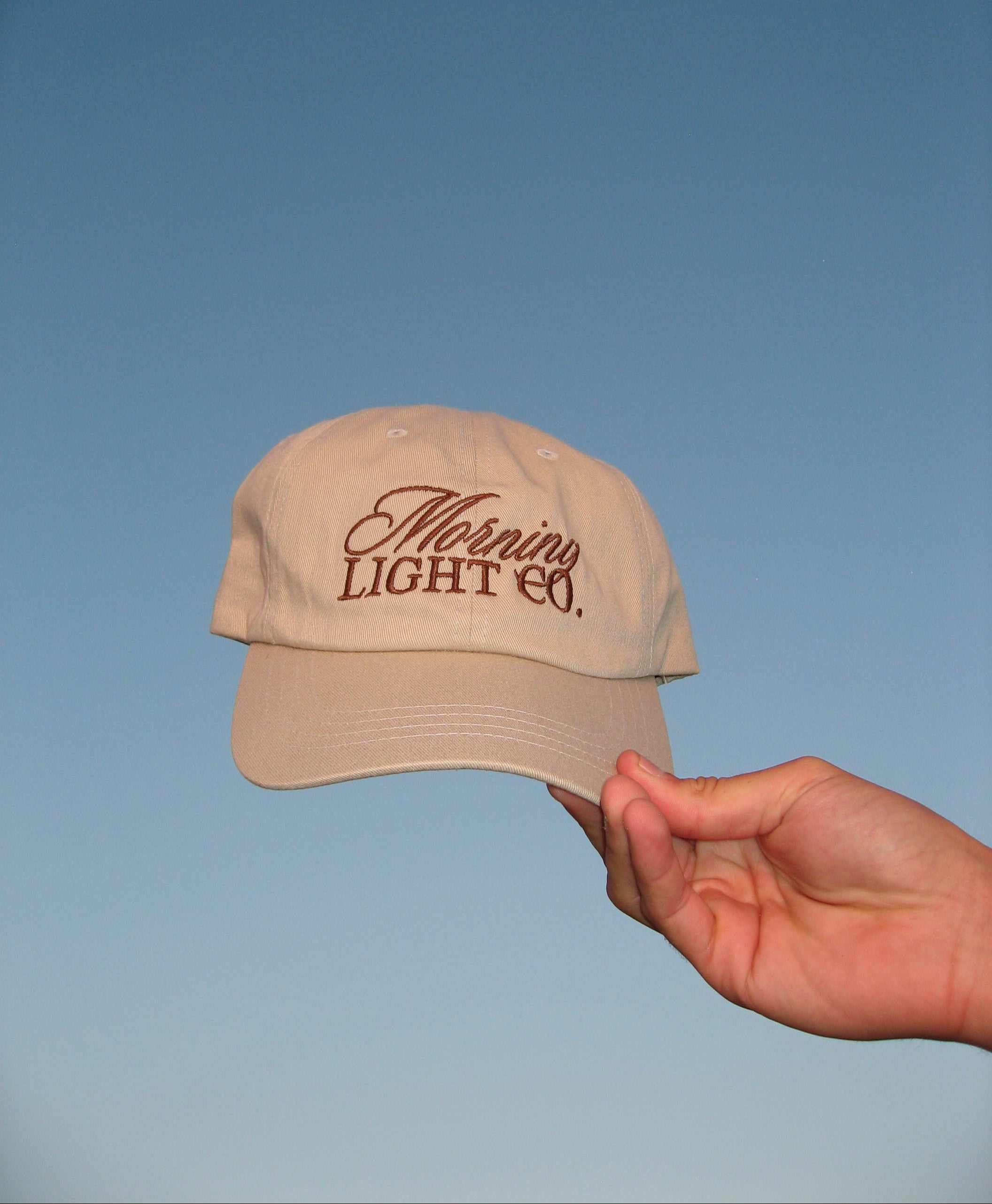 Beige cap with 'Morning Light Co.' text held by a hand against a blue background