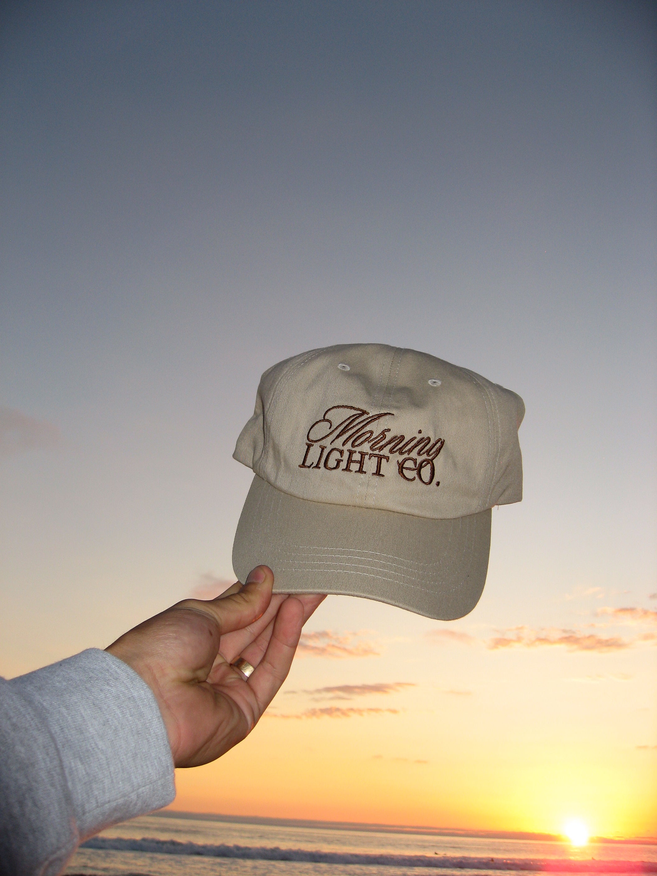 Beige cap with 'Morning Light Co.' text held against a sunset sky