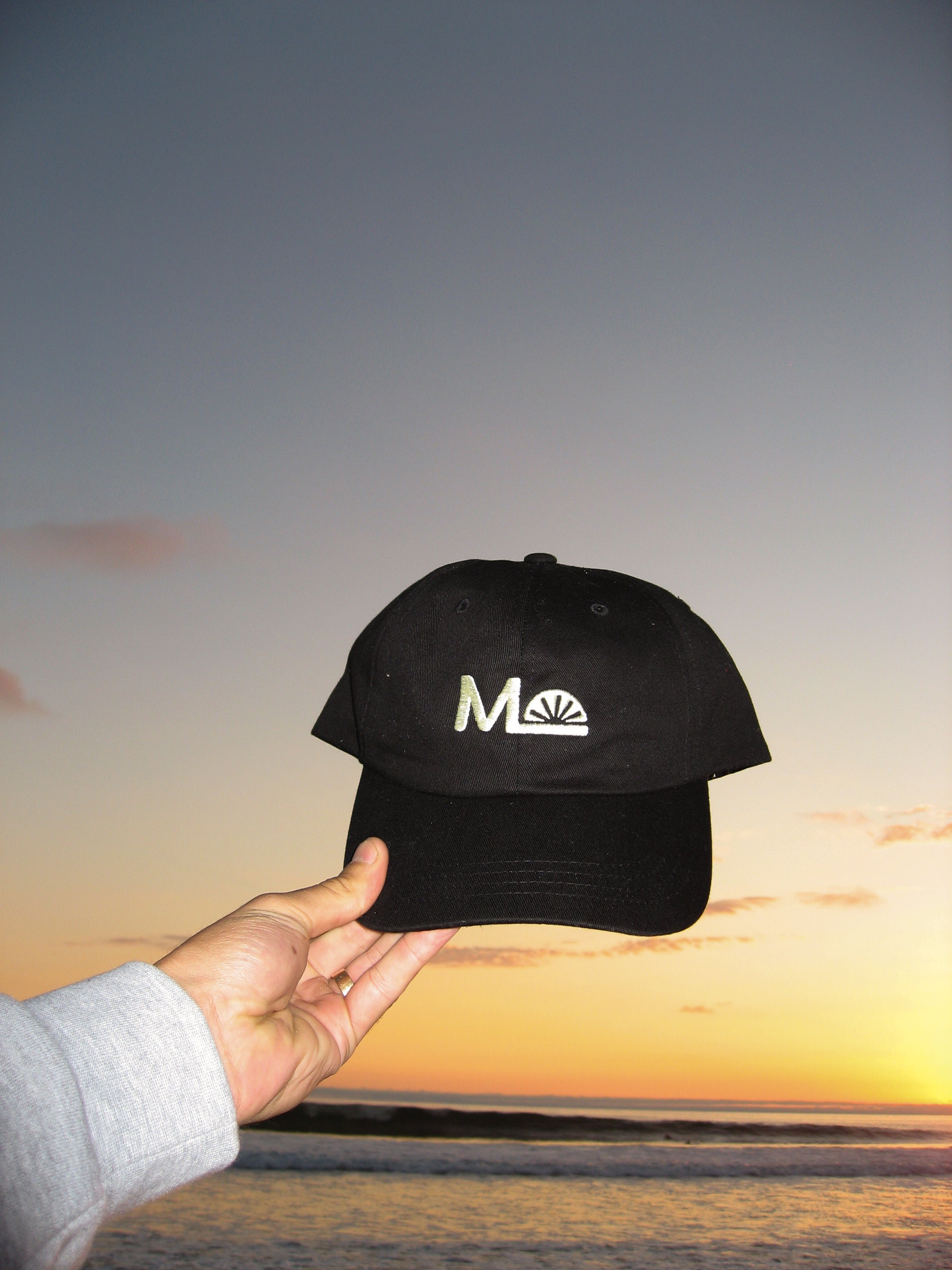 Black cap with a logo held by a hand against a sunset sky.