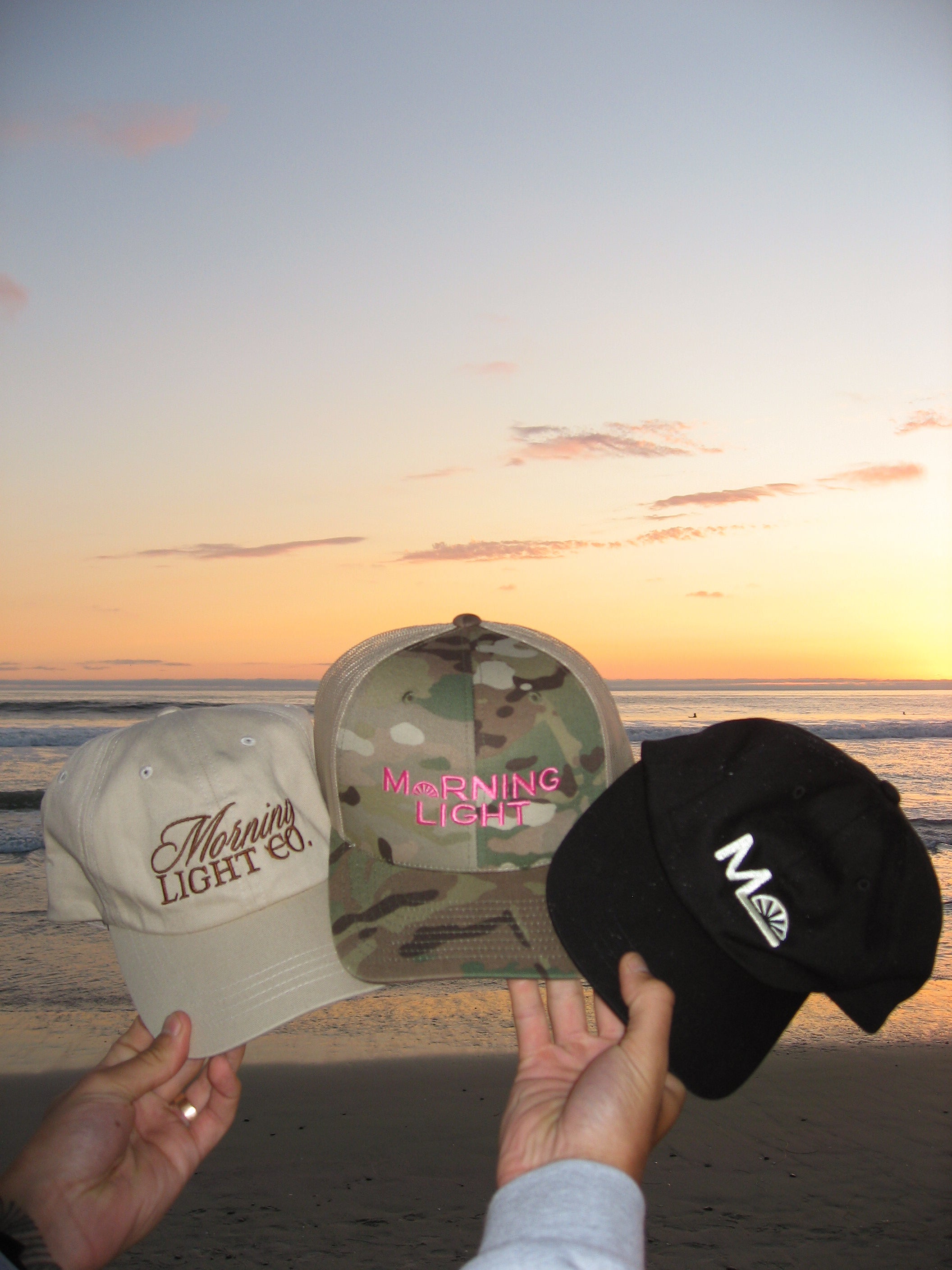 Three hats with text held up against a sunset beach backdrop