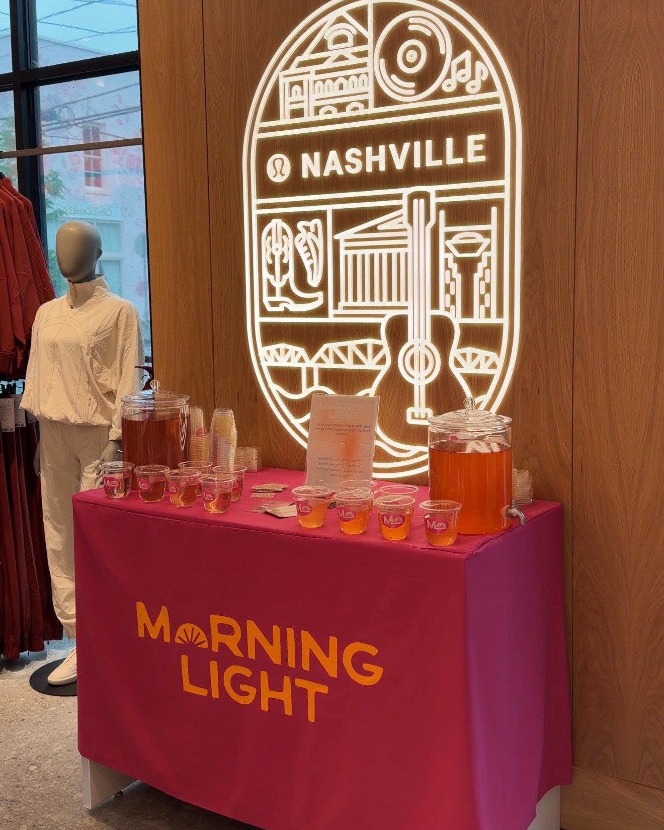 A beverage station with a magenta tablecloth printed with 'MORNING LIGHT' in orange, set against a wood-paneled wall with a large illuminated Nashville graphic.