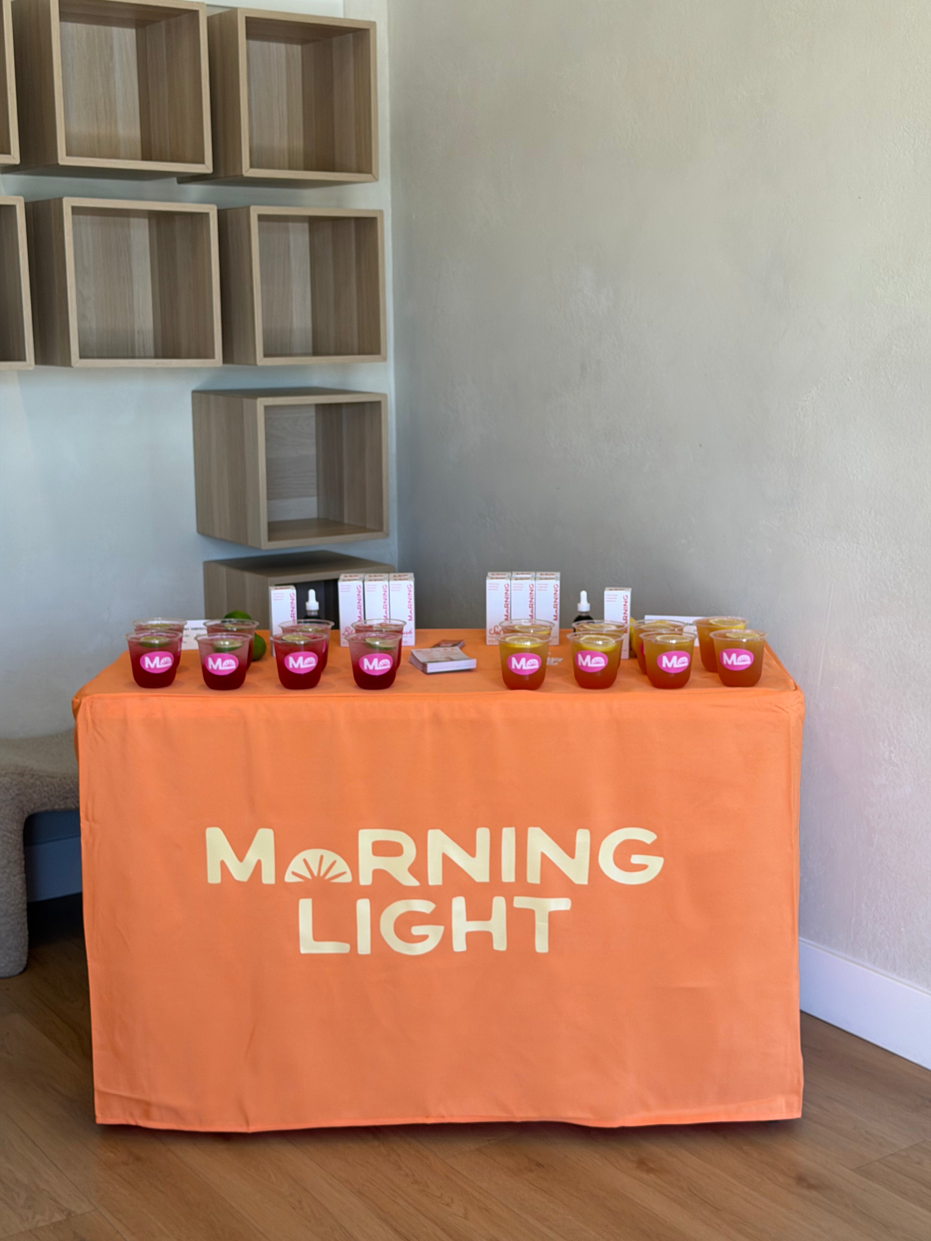 An orange tablecloth with 'MORNING LIGHT' printed in white on the front, draped over a table displaying several pink and amber glass jars and small boxes in an indoor room with wood flooring and wall shelves.