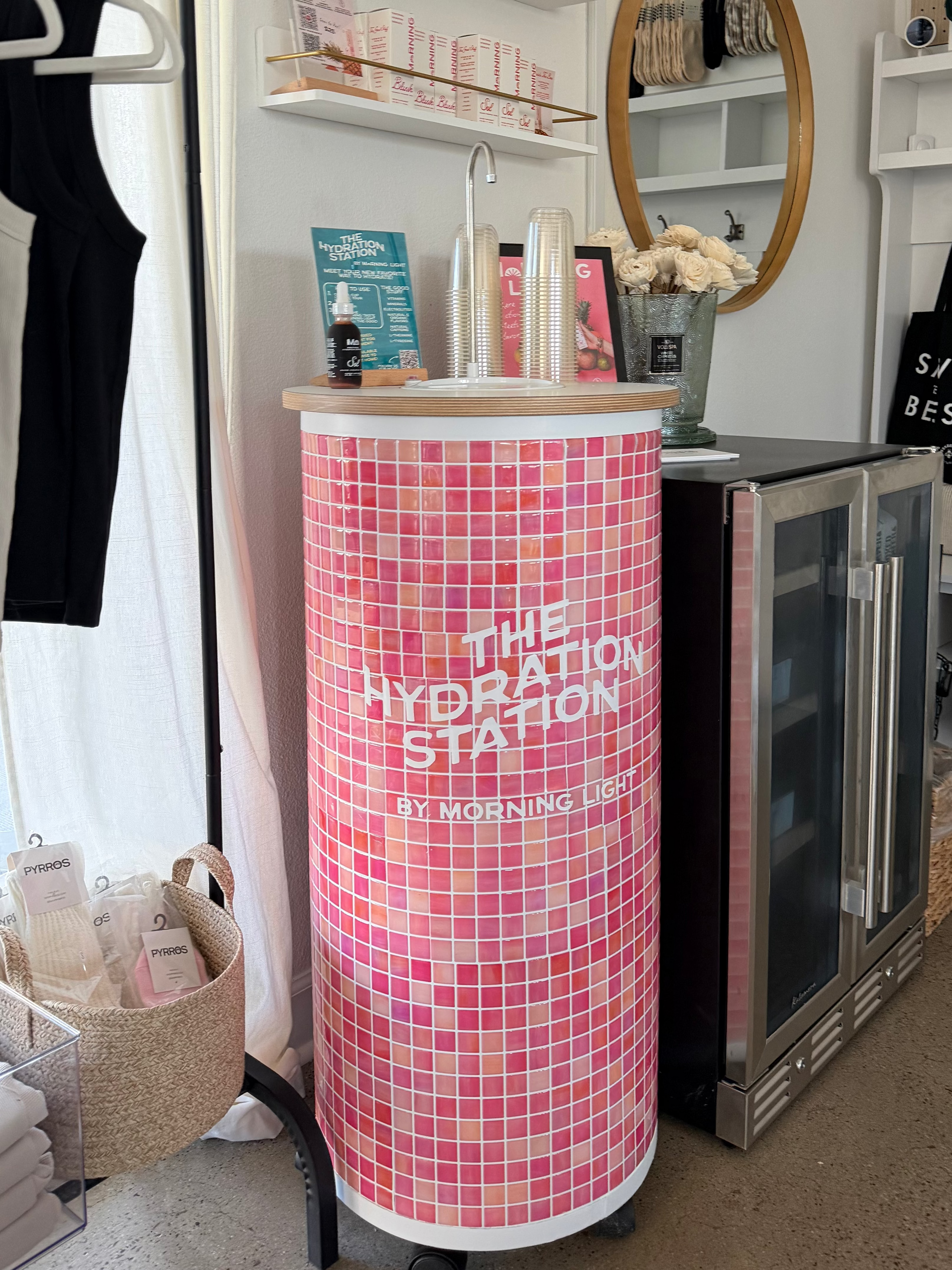 A pink and white mosaic-patterned cylindrical water bottle cooler with the text 'THE HYDRATION STATION' on the front, situated in a retail or salon environment next to a stainless steel cabinet.